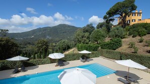 Una piscina con sillones de descanso y sombrillas, rodeada de vegetación y con montañas al fondo.