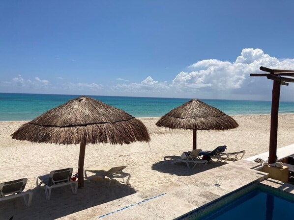A beach with two thatched umbrellas, lounge chairs, and a pool.