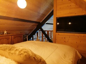 A cozy attic bedroom with a wooden ceiling, a large bed with a fluffy blanket, a window, and a staircase leading to an upper level.