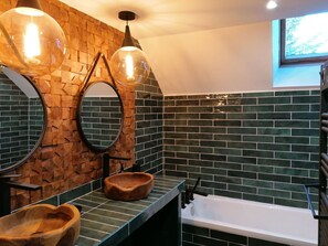 A bathroom with a wooden wall, green tiles, a round mirror, a wooden bowl sink, and a white bathtub.