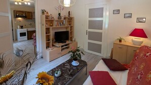 A living room with a TV, shelves, a red lamp, and a door leading to a kitchen.