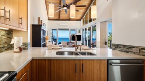 A modern kitchen with wooden cabinets, a stainless steel sink, and a view of the ocean. - 🏖️Oceanfront Penthouse w/ AC + Pickleball + Views (Kailua Kona)