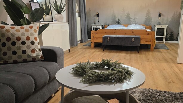 Un salon moderne avec un canapé, une table basse ronde et un lit orné d’une fresque représentant une forêt.