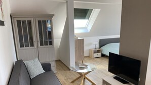 Espaço compacto com sofá, televisão, mesa de vidro, pequena mesa de cabeceira e guarda-roupa com portas de vidro.