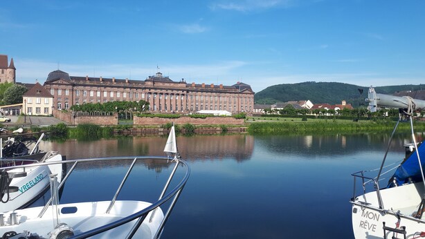 Um rio calmo com barcos atracados, um grande edifício histórico e uma colina ao fundo.
