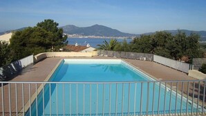 A rectangular swimming pool with a metal fence, surrounded by a wooden deck and trees, overlooking a coastal landscape with mountains in the distance.