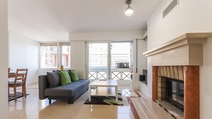 A spacious living room with a sofa, a coffee table, and a fireplace. There is a balcony with a railing and a door leading outside. - Lisbon Flower 360º - Your Lovely Flat with Pool and Parking (Lisboa)