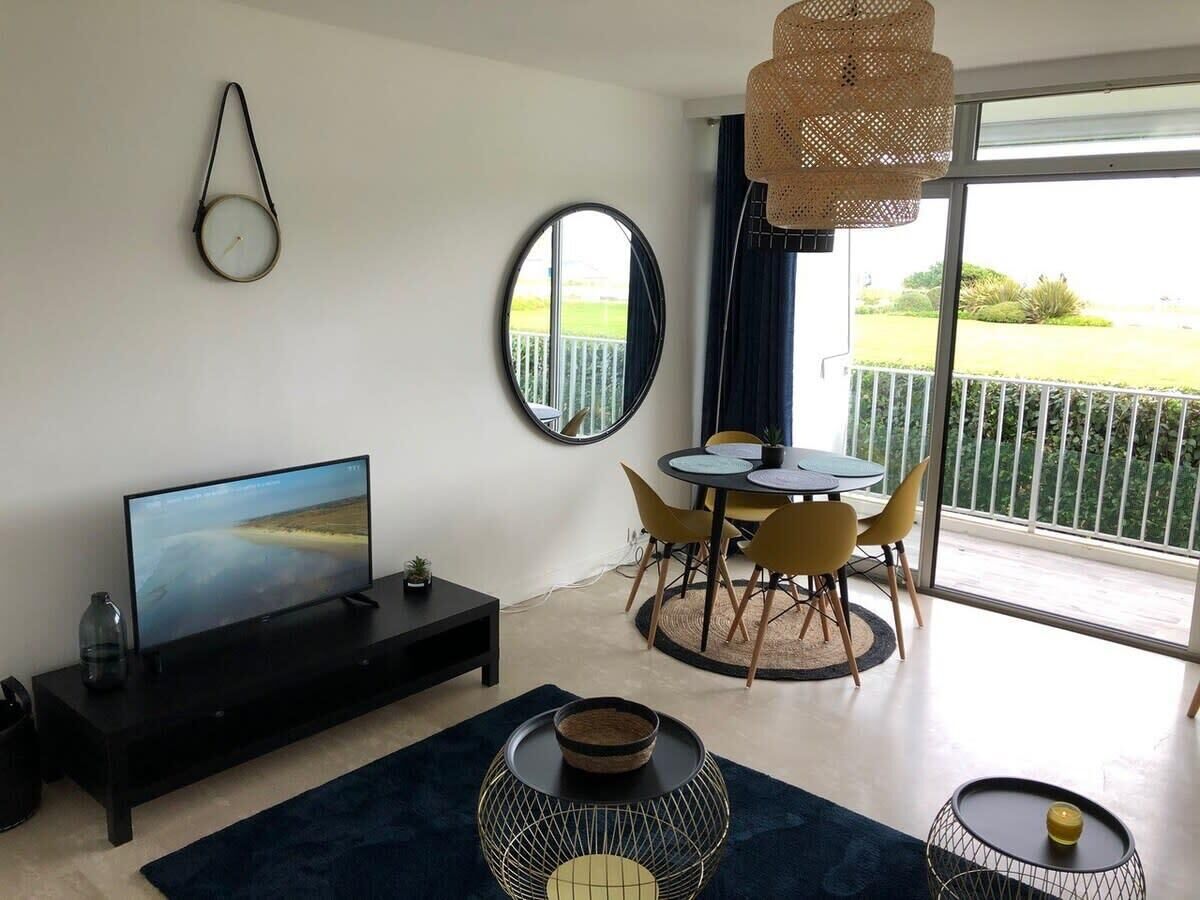 A modern living room with a flat-screen TV, a dining area with yellow chairs, a round table, and a balcony with a view.