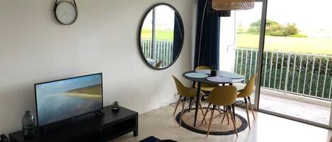 A modern living room with a flat-screen TV, a dining area with yellow chairs, a round table, and a balcony with a view.