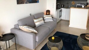 Een moderne woonkamer met een grijze bank, een klein zwart bijzettafeltje en een donkerblauwe vloerkleed. Achtergrond is een keuken zichtbaar met witte kasten en een magnetron.