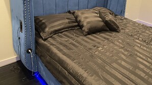 A room with a blue upholstered headboard, a bed with dark striped bedding, and a blue under-bed lighting strip.