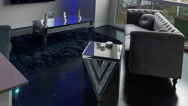 A modern living room with a flat-screen TV mounted on the wall, a glass coffee table, a dark blue rug, a grey sofa, and a large window offering a view of buildings outside.