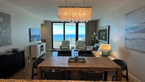 A modern hotel room with a dining area, a large chandelier, a television, and a view of the ocean. - Gulf Front Condo with Stunning Views! (Pensacola)