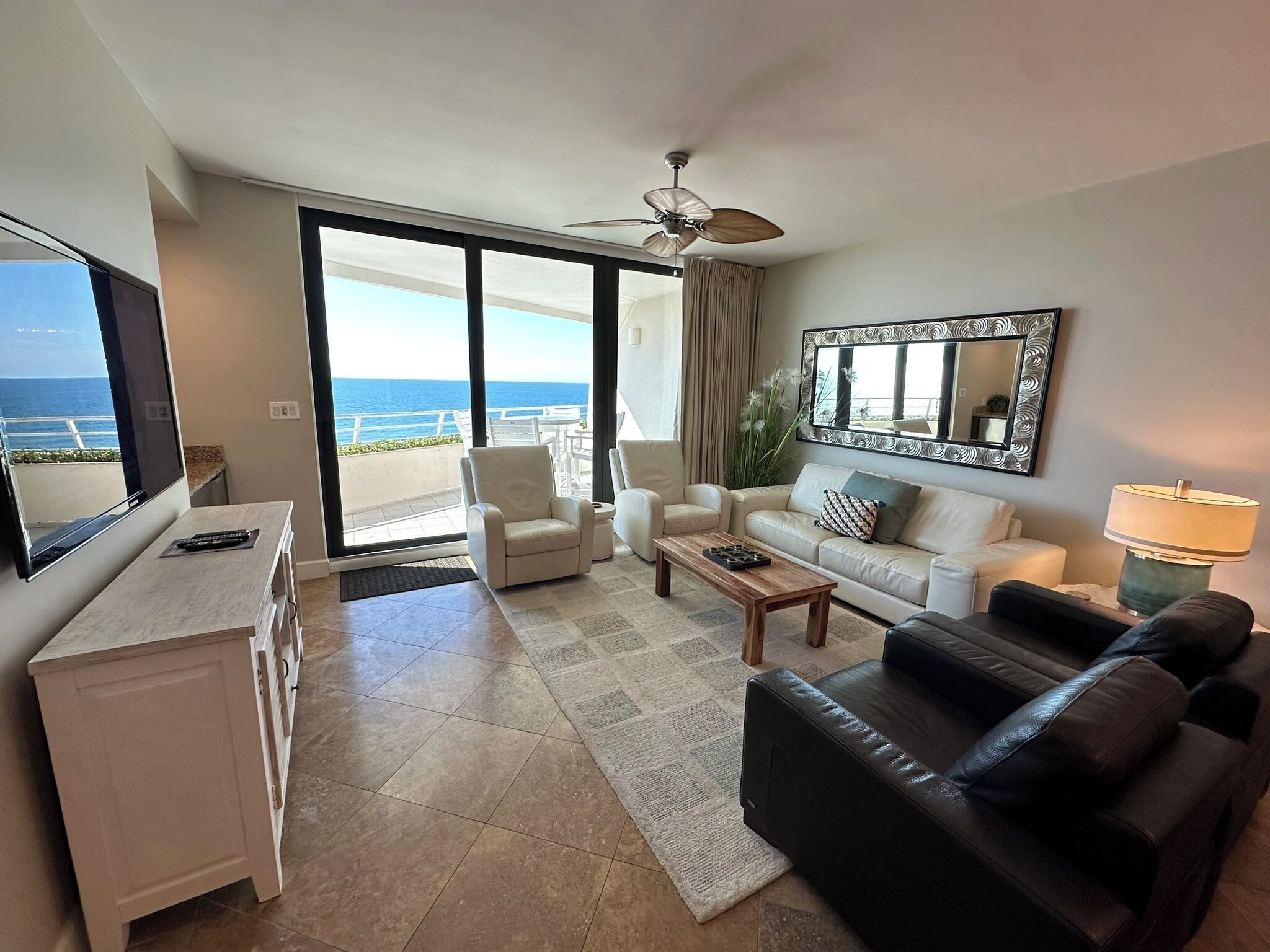 A modern living room with a large TV, a ceiling fan, and a view of the ocean.