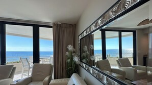 A modern hotel room with a large window offering a view of the ocean. There is a beige sofa, a white armchair, and a large mirror with an ornate frame. - Gulf Front Condo with Stunning Views! (Pensacola)