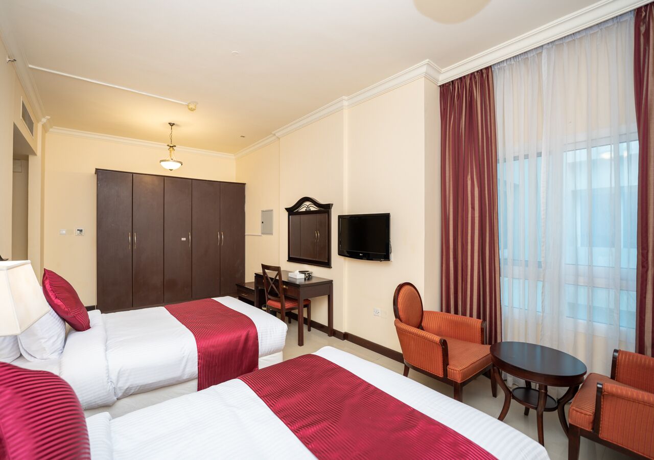 Ein Hotelzimmer mit zwei Betten, einem Schreibtisch, einem Sessel, einem Fernseher und einem großen Fenster mit Vorhängen.