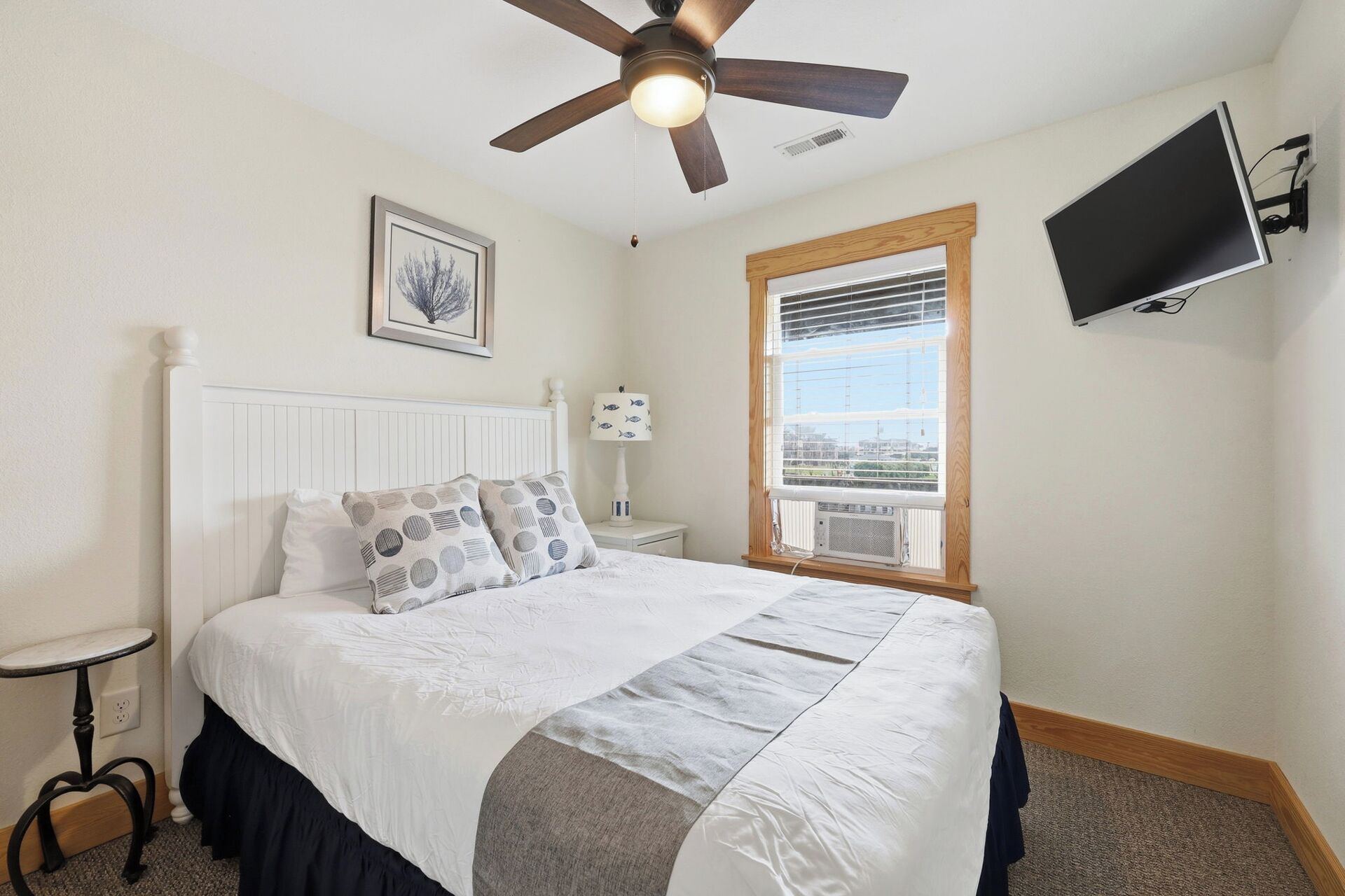 Une chambre à coucher comprenant un lit, un ventilateur de plafond, un téléviseur fixé au mur, une fenêtre avec des stores et un tableau encadré accroché au mur.