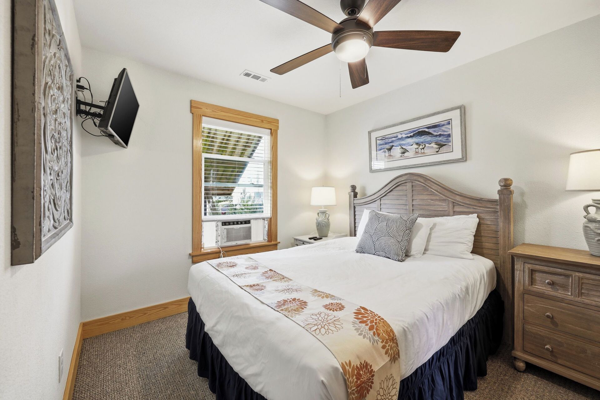 Une chambre à coucher comprenant un lit, un ventilateur de plafond, une fenêtre avec des stores, une table de chevet avec une lampe et une commode.