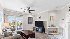 A living room with a sofa, coffee table, TV, and a balcony with a view of buildings. - Salty But Sweet at Sundune Village, Located 0.1 Miles from the Beach! (Kill Devil Hills)