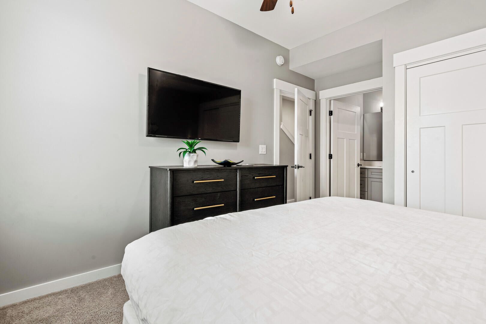 A bedroom with a large bed, a dark dresser with drawers, a flat-screen TV mounted on the wall, and a door leading to a bathroom.
