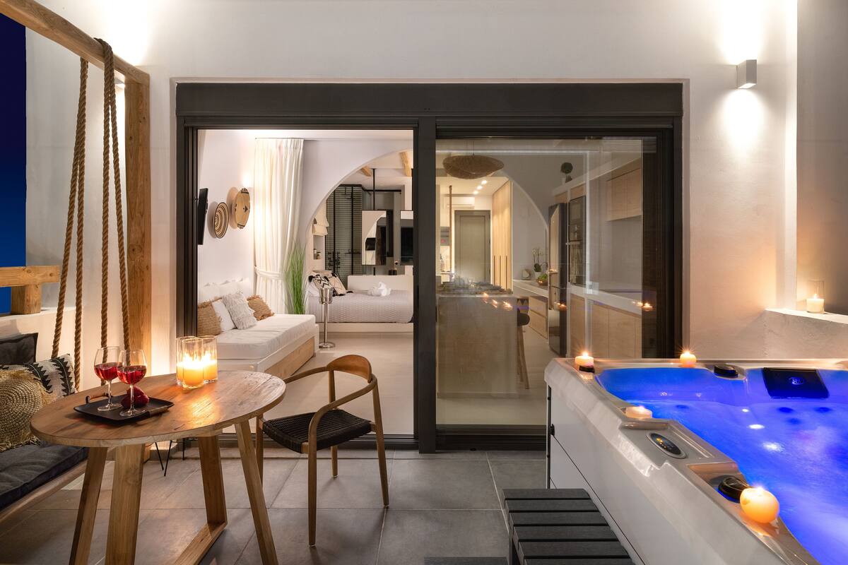 A modern room with a glass door leading to a living area, a wooden table with glasses and a candle, and a hot tub with candles.