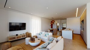 A modern living room with a large TV, white sofas, a coffee table, and a kitchen area in the background. - Periyiali Resort , Villa Melpomeni 21 at Konnos Beach - Cape Greco (Paralimni-Protaras)