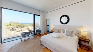 A modern bedroom with a large bed, a view of the landscape, and a seating area with a table and chairs. - Periyiali Resort , Villa Melpomeni 21 at Konnos Beach - Cape Greco (Paralimni-Protaras)