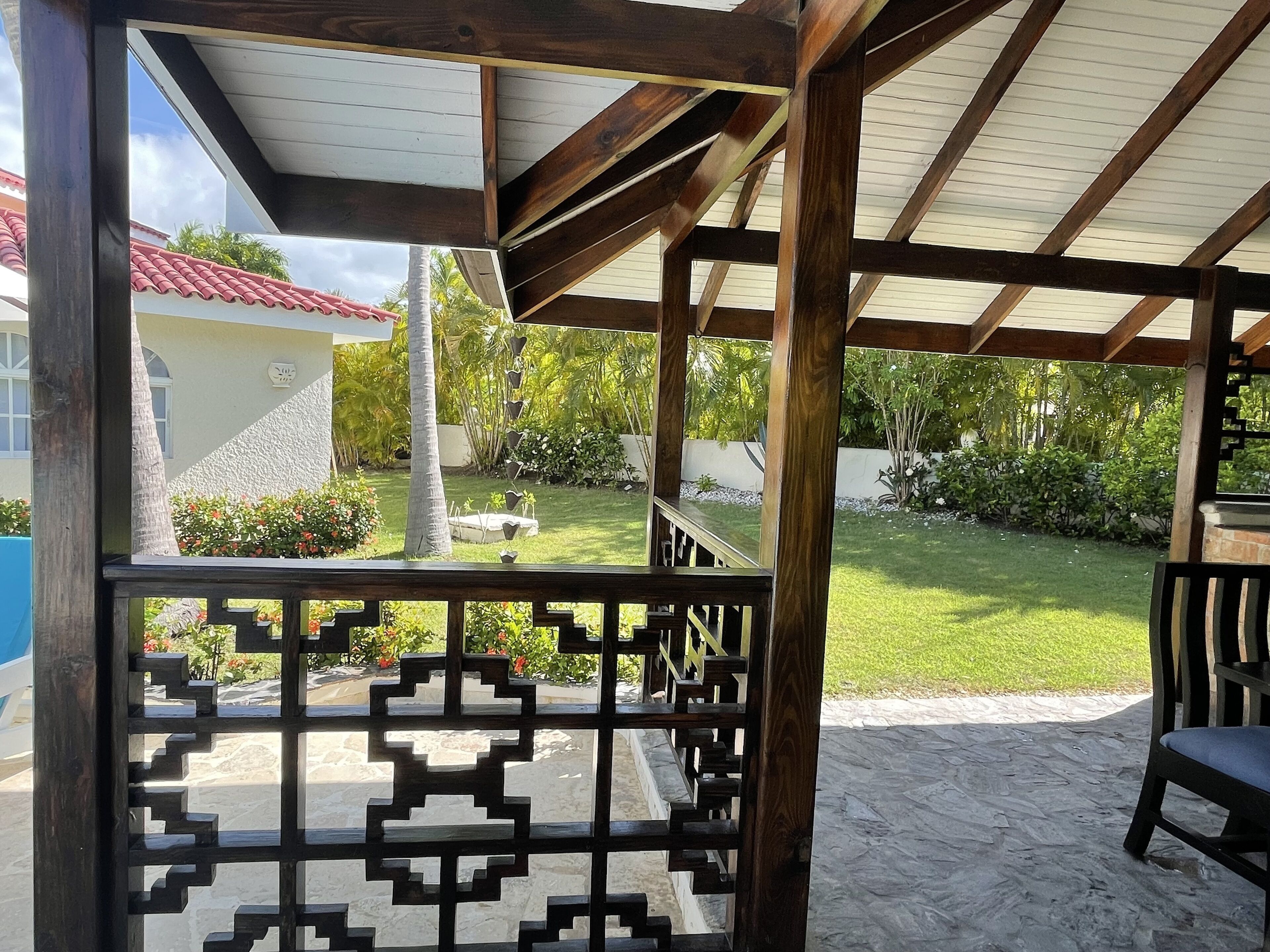 Un espace extérieur abrité, doté d’une pergola en bois, d’un sol en pierre et offrant une vue sur un jardin avec, en arrière-plan, un bâtiment blanc et un toit recouvert de tuiles rouges.