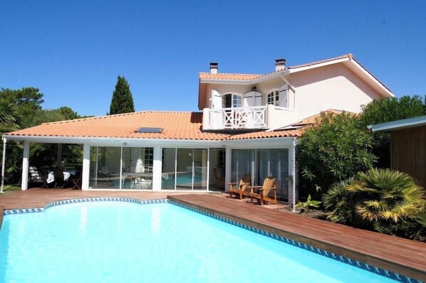 Una piscina con un porche cubierto, una casa de dos pisos con balcón y un cielo azul despejado.