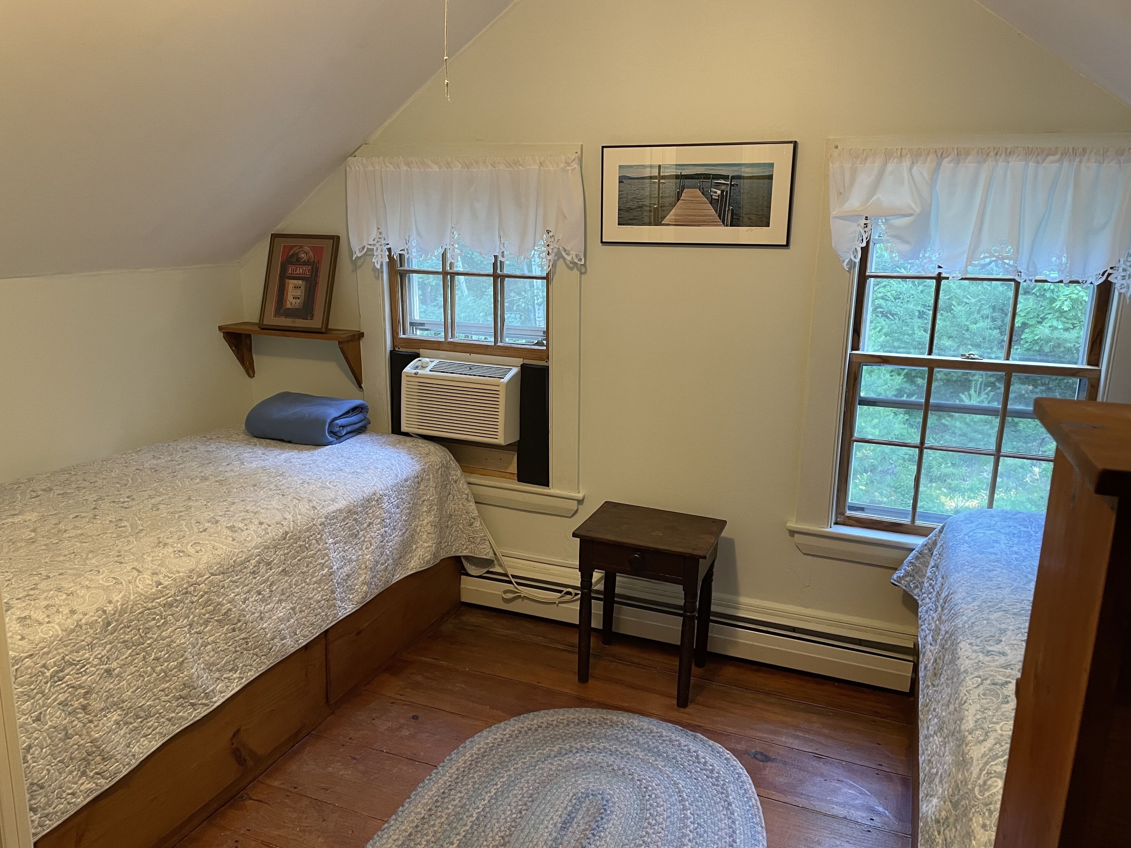 A bedroom with two beds, a window with a view of greenery, a framed picture on the wall, and a small table.