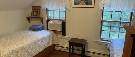 A bedroom with two beds, a window with a view of greenery, a framed picture on the wall, and a small table.