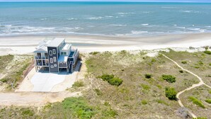 A modern beach house with a large deck and multiple windows, situated on a sandy beach with a view of the ocean.