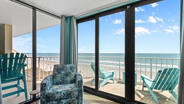 A balcony with a view of the beach, featuring a patterned armchair and two green Adirondack chairs. - Crescent Sands In Windy Hill D6 3 Bedroom Condo by RedAwning (North Myrtle Beach)
