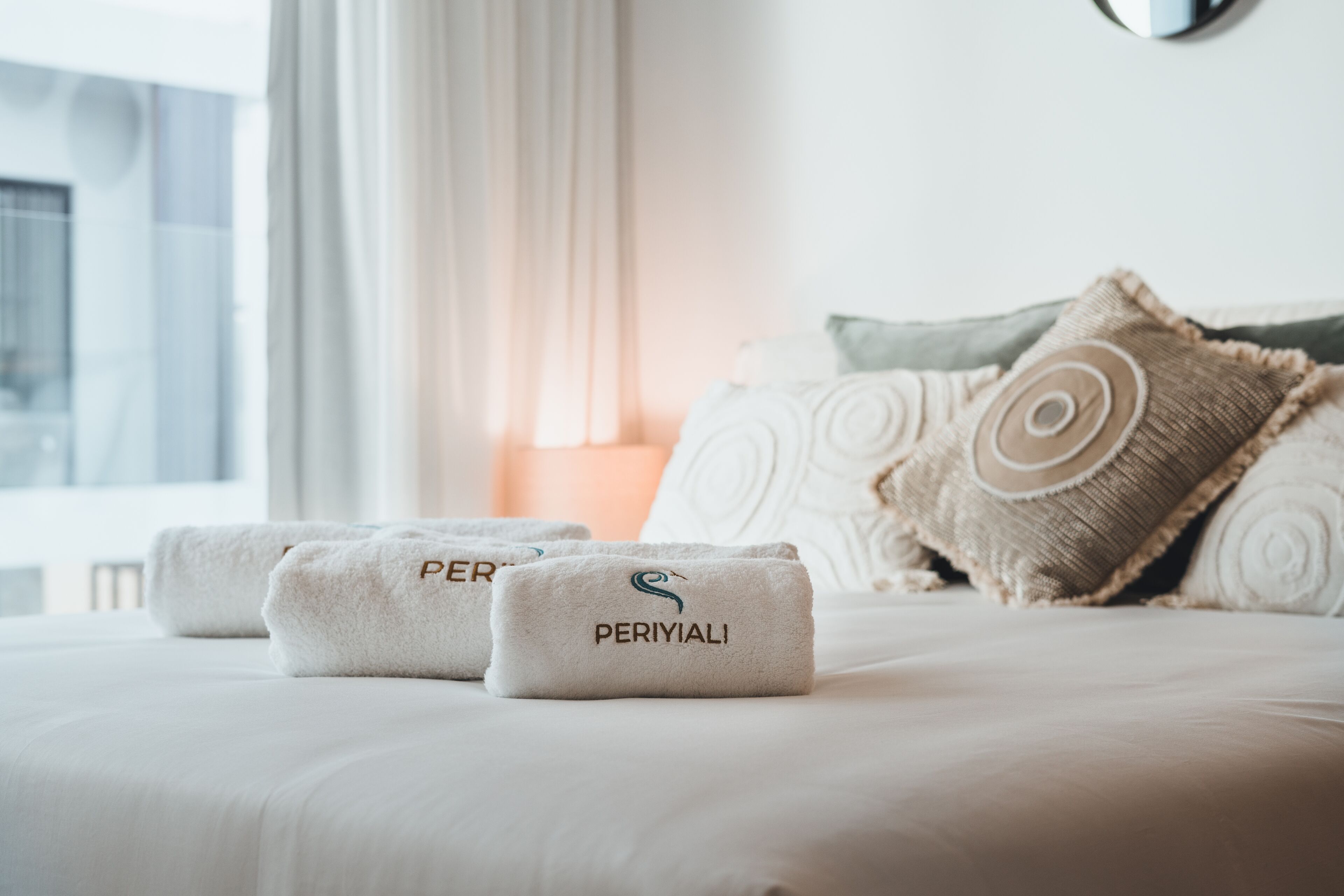 A neatly made bed with two towels labeled "PERIYALI" and a decorative pillow.
