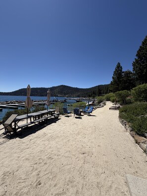 A beach with lounge chairs and umbrellas, a clear sky, and a lake with a bridge in the background. - Magnificent 2-bedroom, 2-bath lakefront complex (Incline Village)