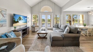 A spacious living room with a large TV, comfortable seating, and a view of the outdoors through large windows.