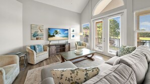 A spacious living room with a high ceiling, a large window with blinds, a flat-screen TV, and a cozy seating area with a coffee table.