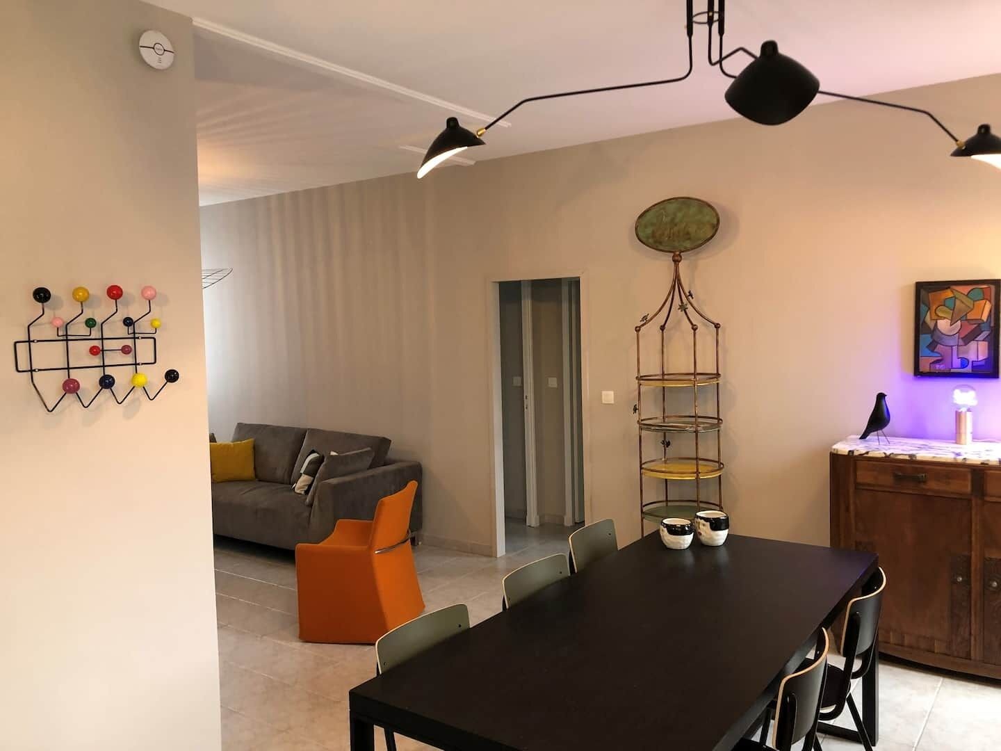 Ett modernt vardagsrum med ett matbord, en soffa och ett soffbord. Det finns en väggmonterad lampa och ett dekorativt metallställ.