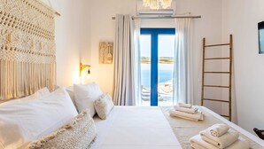 A bedroom with a large bed, a view of the sea through a blue door, a macrame wall hanging, and a wooden ladder.