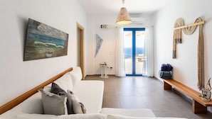 En moderne stue med en hvid sofa, et maleri af en strandscene, en træbænk og havudsigt gennem en glasdør.