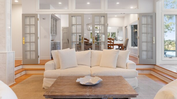 A spacious living room with a white sectional sofa, a wooden coffee table, and a view of the kitchen through glass doors. - Sunset Marsh Estate/8BR/Horizon pool/Renovated Gourmet kitchen/sleeps 18 (Kiawah Island)