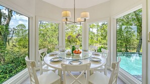 A round dining table with white chairs, a chandelier, and a view of a swimming pool and greenery. - Sunset Marsh Estate/8BR/Horizon pool/Renovated Gourmet kitchen/sleeps 18 (Kiawah Island)