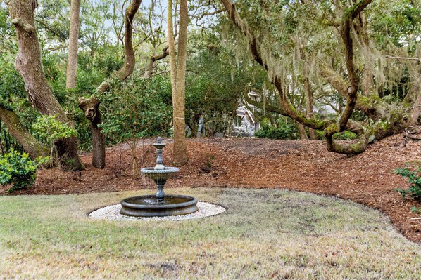 A garden with a central fountain, surrounded by trees and a white building in the background. - Brand new renovation, right across the street from the beach, private pool (Kiawah Island)
