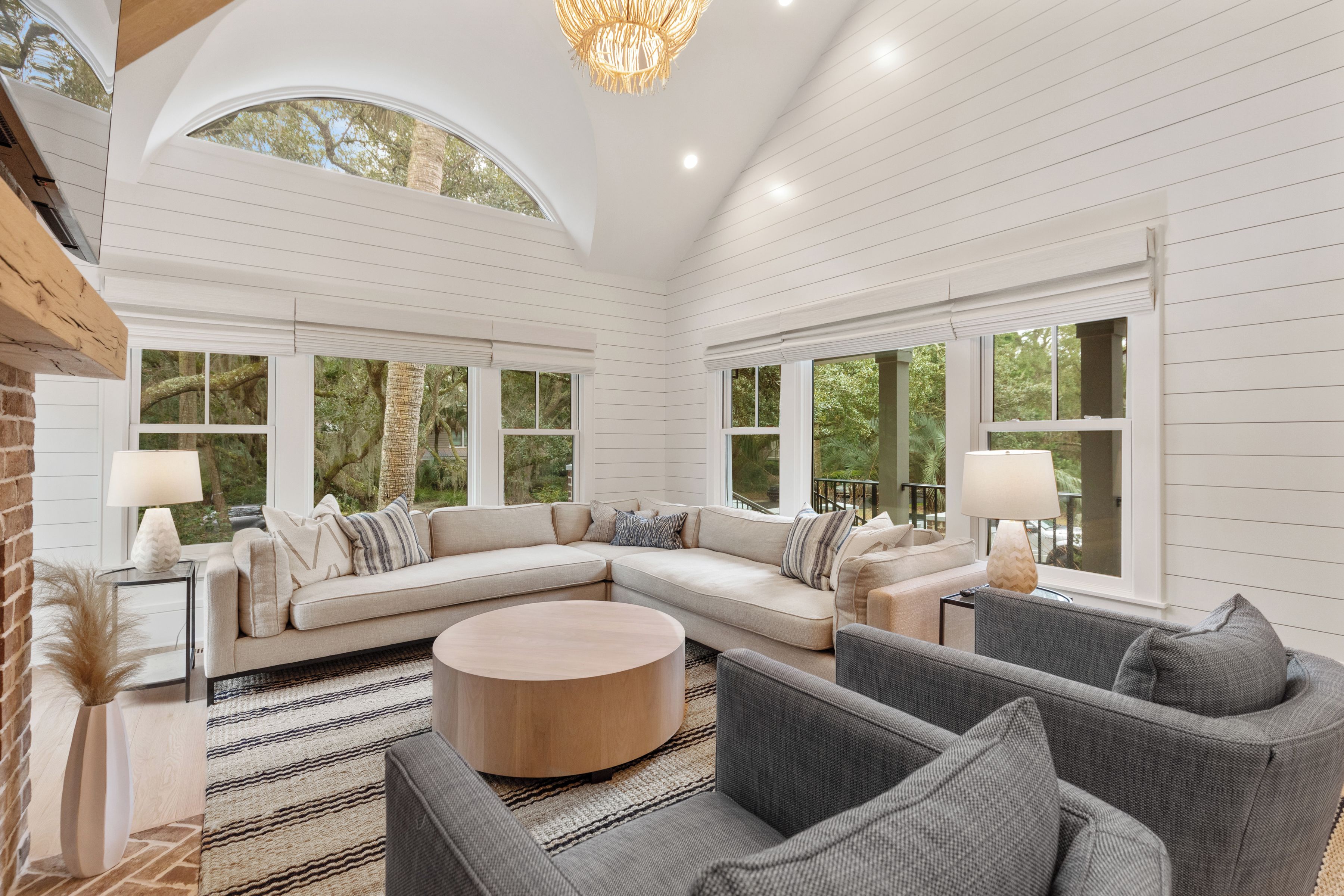 A spacious living room with a large L-shaped sectional sofa, a round coffee table, and a gray couch. There are two side tables with lamps, and the room features white paneled walls and large windows.