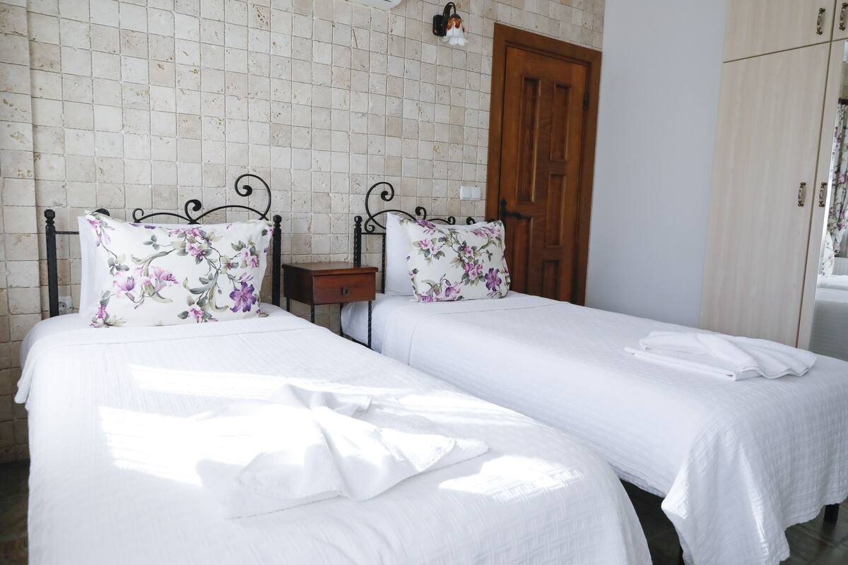A room with two beds, each with floral-patterned pillows and white linens. There is a wooden door and a wardrobe in the background.