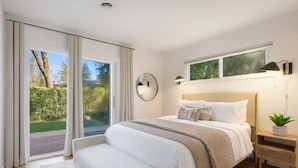A modern bedroom with a large bed, a bedside table with a lamp, a small plant, and a view of a garden through the sliding glass door.
