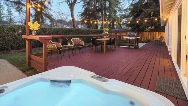 A hot tub on a wooden deck with string lights, outdoor furniture, and a barbecue grill.