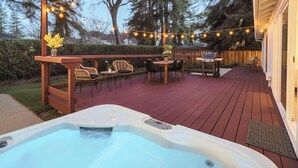 Un bain à remous sur une terrasse en bois, agrémentée de guirlandes lumineuses, de meubles d’extérieur et d’un barbecue.