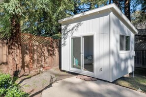 Ein kleines, weißes, eingeschossiges Gebäude mit Schiebeglastür, einem Fenster und einer Holzterrasse.