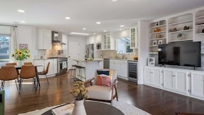 A modern kitchen with white cabinetry, a dining area with a table and chairs, and a living room with a TV and shelves.